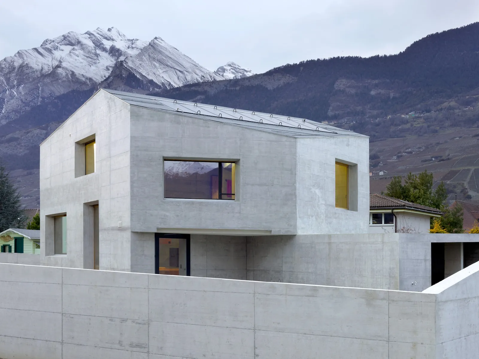Maison contemporaine en béton brut, toit à deux pans, au pied de montagnes enneigées